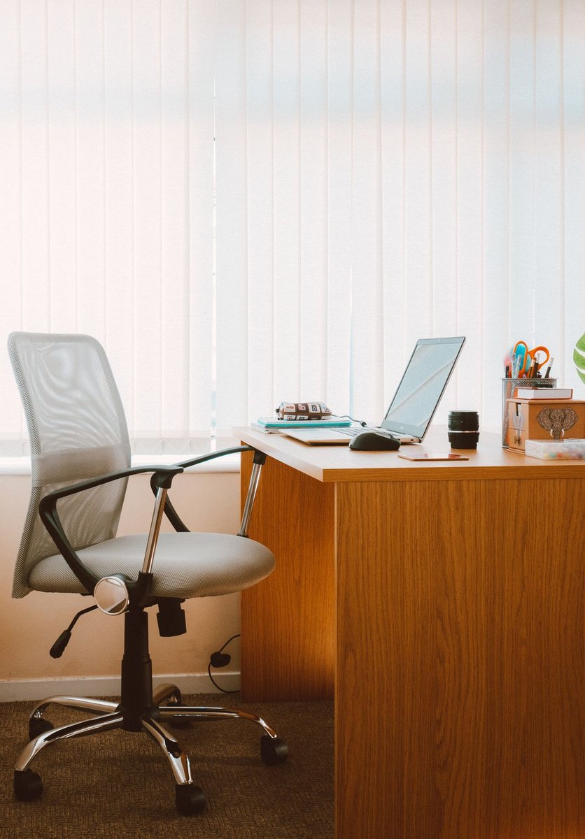 Grey mesh office chair with armrests beside a wooden desk holding a laptop, in front of window blinds